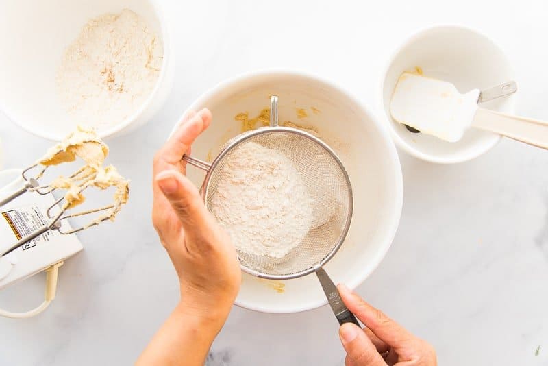 Two hands sift the dry ingredients into a white ceramic bowl.