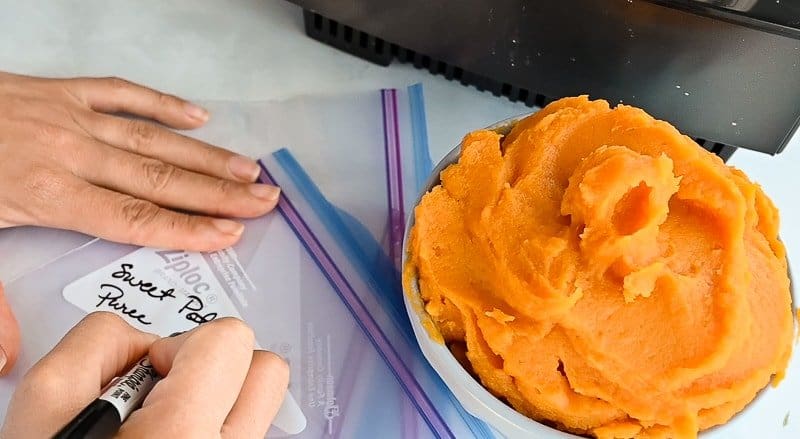 Hand writes the words “sweet potato puree” in black marker on a food storage bag next to a blue bowl filled with sweet potato puree.