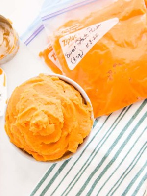 Sweet potato puree in a gray bowl next to a green and white striped kitchen towel bags filled with sweet potato puree are marked with a date