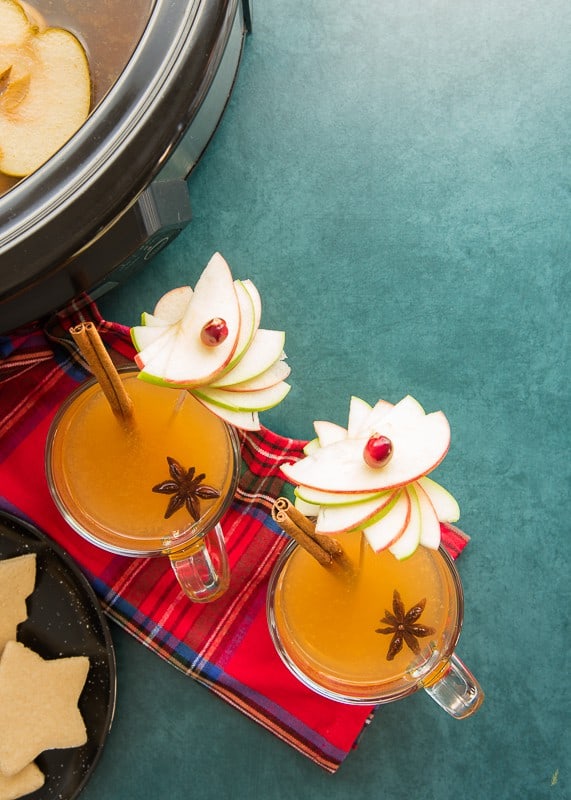 Portrait image of two clear glass mugs filled with slow cooker wassail garnished with apple pinwheels, cinnamon sticks, star anise on a teal green background. A red plaid cloth napkin sits underneath the mugs.