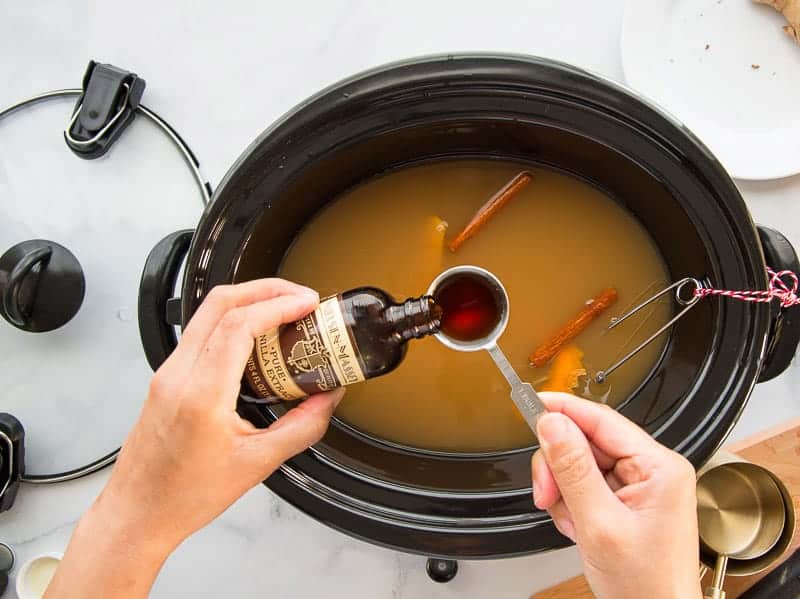 A hand measures out vanilla extract from its bottle Into the slow cooker with the rest of the ingredients.