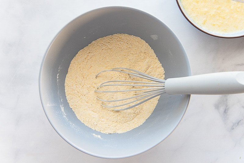 The cornmeal and flour mixture is whisked together with a grey whisk in a large blue mixing bowl