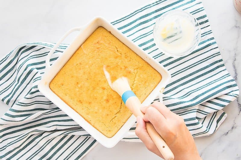 A hand uses a wooden pastry brush to brush melted butter on the baked cornmeal in a white square baking dish.