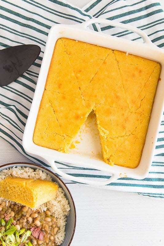A portrait image of the baked cornbread in a white square baking dish on a green and white striped kitchen towel