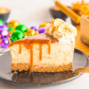 A slice of Bananas Foster Cheesecake drizzled with rum caramel on a black plate in front of gold, green, and purple Mardi Gras beads.