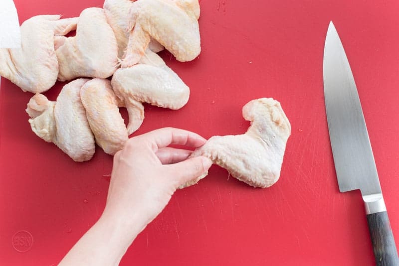 A hand holds a whole raw chicken wings which is to the left of a silver knife on a red cutting board