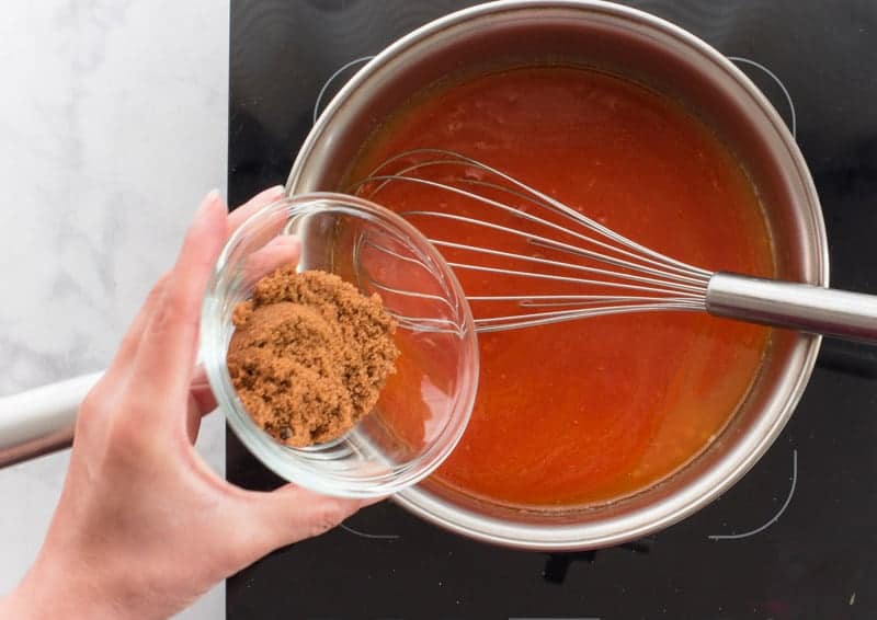 A hand adds brown sugar from a clear glass bowl to a pot of buffalo wing sauce