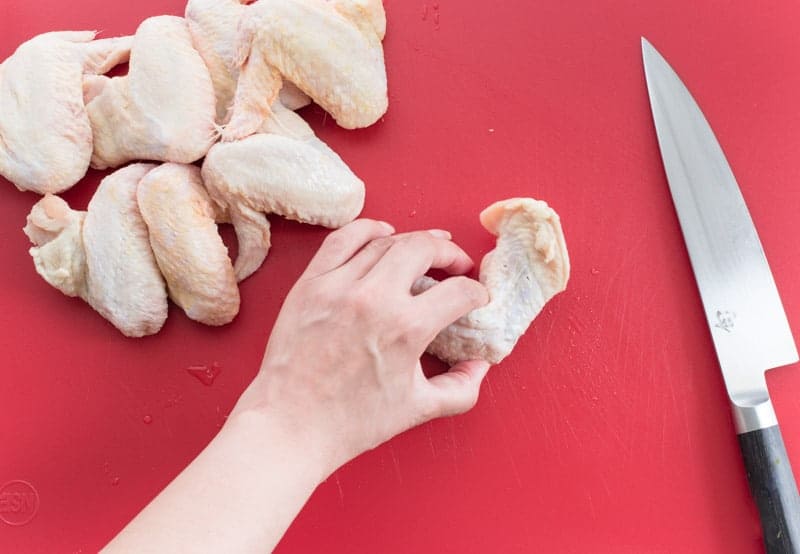 A hand pinches the joint of a chicken wing to indicate where to cut. Knife to the right on a red cutting board