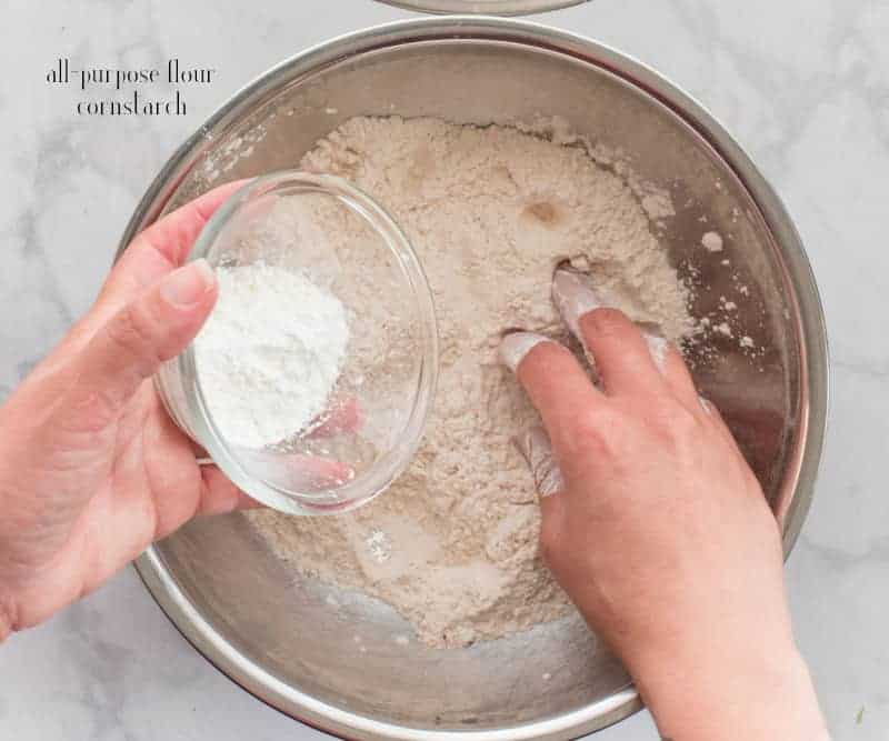 A hand adds cornstarch from a white ramekin to flour in a metal mixing bowl