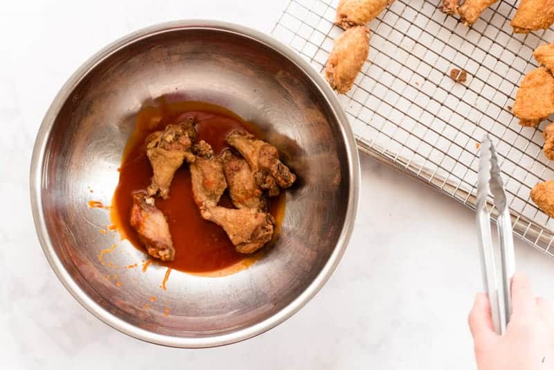 Fried wings added to a silver bowl of buffalo sauce a hand holds metal tongs on right of image.