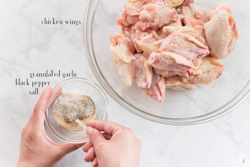 A hand uses a measuring spoon to combine the seasoning for the chicken wings in a clear glass bowl. Top right raw chicken wings in a glass bowl. Black text overlay reads: chicken wings, granulated garlic, black pepper, salt.