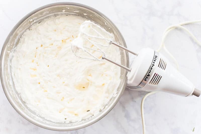 An electric hand mixer's beaters are covered in whipped cream above a bowlful of whipped cream