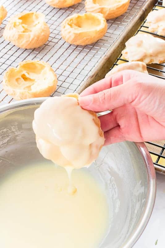 A hand removes the top of a pate a choux shell from a bowlful of white chocolate ganache after dipping it