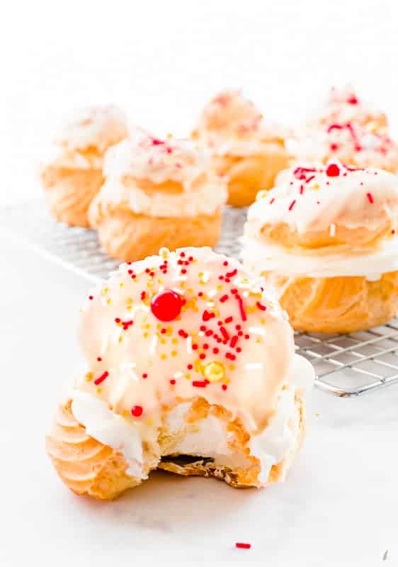A cream puff with a bite removed sits in front of a cooling rack that has more cream puffs on it.