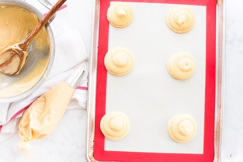 The pate a choux paste is piped onto a red and white silicone baking mat-lined cookie sheet.
