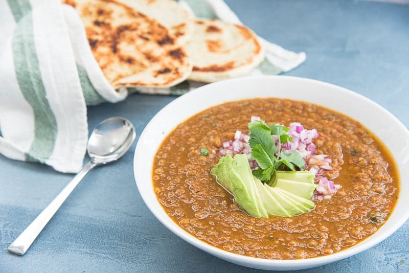 three-quarter image Vegan Lentil Stew in a white bowl garnished with avocado and red onion silver spoon blue surface green and white towel wrapped around naan.