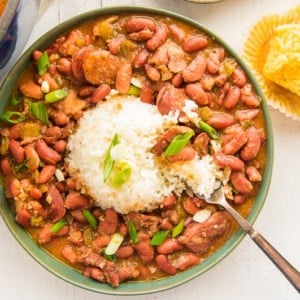 Overhead image of a green bowl of creole red beans and rice with a silver fork. Garnished with green onion. An unwrapped cornbread muffin with a bite removed on top right.