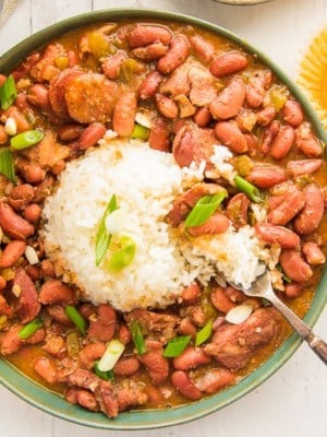 Overhead image of a green bowl of creole red beans and rice with a silver fork. Garnished with green onion. An unwrapped cornbread muffin with a bite removed on top right.