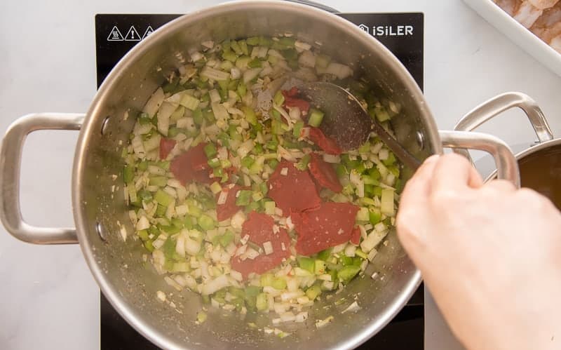 Tomato paste is added to green peppers, onions, celery, and garlic in a silver pot.