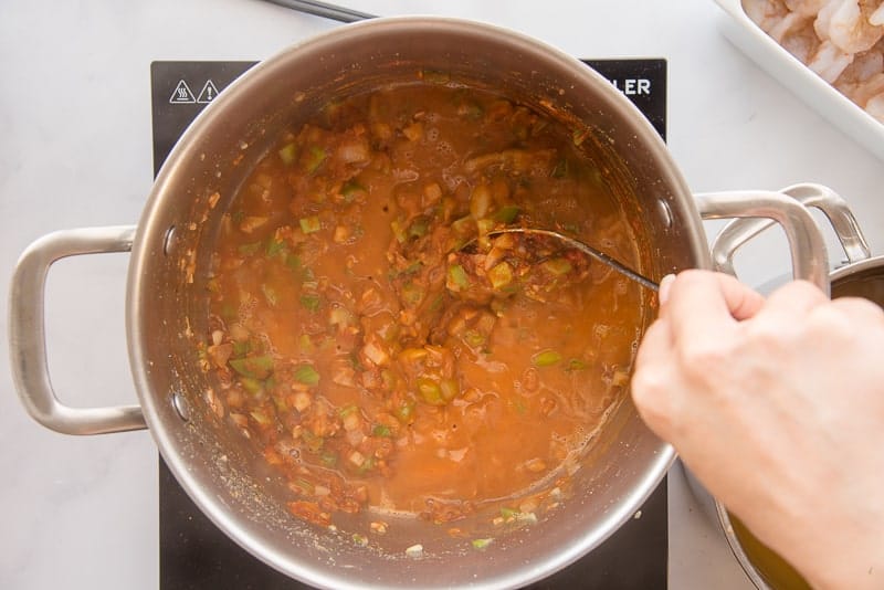 A hand uses a silver spoon to stir the stock into the roux in a silver pot.