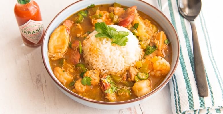 Close-up horizontal image blue bowl white rice gumbo, green and white striped napkin, black spoon on top, tabasco bottle