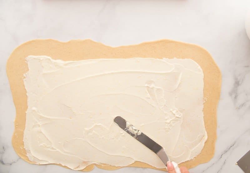 A spatula spreads cream cheese filling on a rectangle of dough.