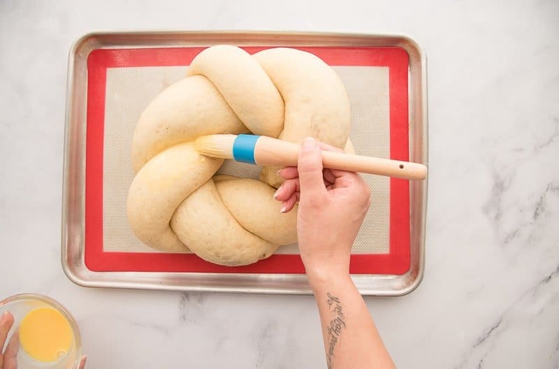 A hand uses a pastry brush to glaze unbaked King Cake in egg wash
