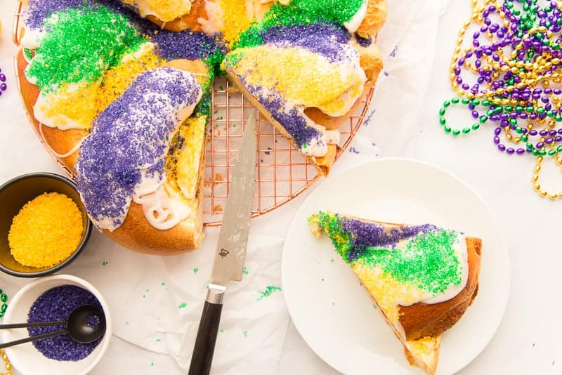 Horizontal image of a whole King Cake with a slice removed. The slice is on a white plate surrounded by sanding sugar and Mardi Gras beads