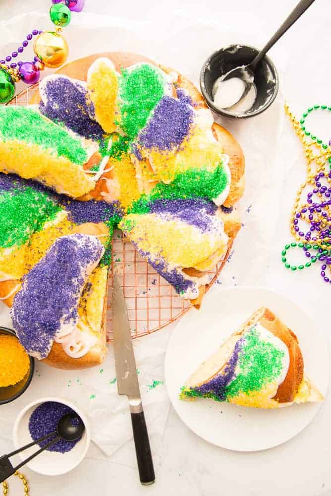 A King Cake decorated in purple, yellow, and green sanding sugar. A slice is removed and on a white plate to the bottom right.
