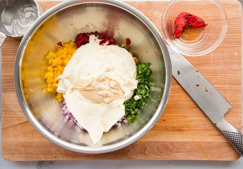 Mayonnaise and mustard added to chopped veggies and herbs in a silver mixing bowl on a wooden cutting board.