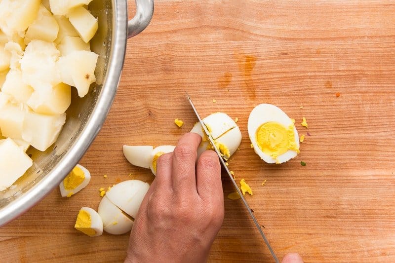 Hands holding a boiled egg and a knife used to chop them on a wooden cutting board.