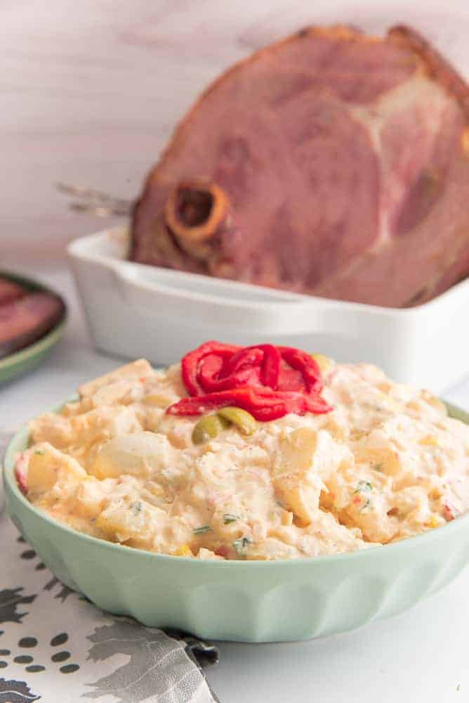 A green serving bowl of Puerto Rican Potato Salad. Red pepper garnish on top. Top left of image a white roasting pan with a ham.