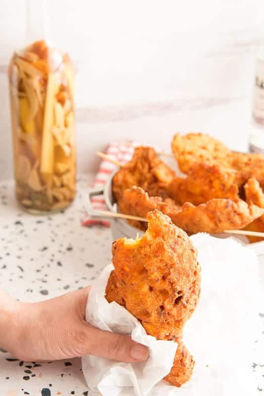 Portrait image hand holding a bacalaito wrapped in white paper in front of a silver pot of bacalaitos on skewers and a bottle of hot sauce in the background