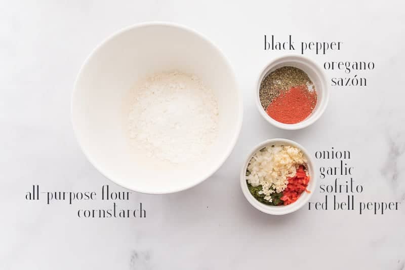 Ingredients to make bacalaitos on a white countertop: flour, cornstarch, pepper, oregano, sazón, onion, garlic, sofrito, and red bell pepper
