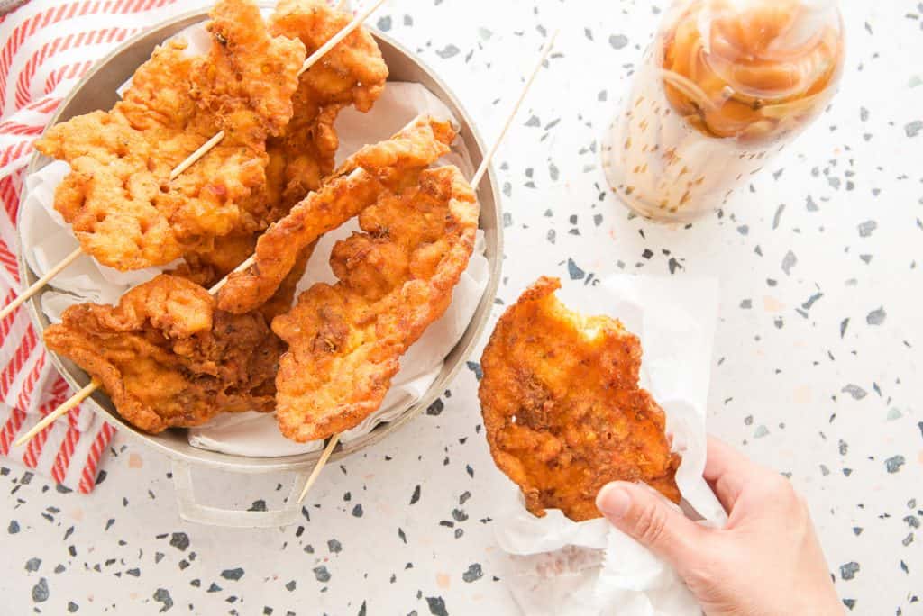 Preview image a hand holding a codfish fritter wrapped in white wax paper over a terrazzo surface next to a silver pot of fritters on wooden skewers.