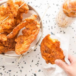 Preview image a hand holding a codfish fritter wrapped in white wax paper over a terrazzo surface next to a silver pot of fritters on wooden skewers.