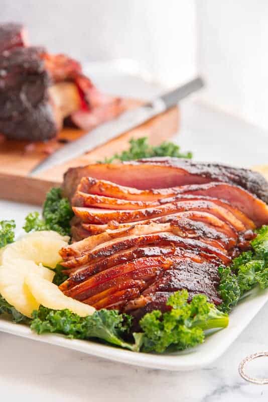 Portrait image of a white platter with a bed of greens and sliced spiced brown sugar pineapple ham on it.