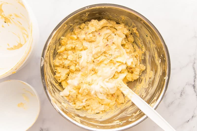 Mayonnaise is folded into the potato salad in a silver bowl with a large white spatula