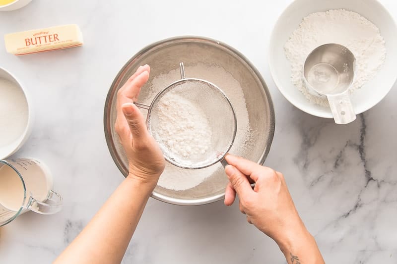 Flour, ground ginger, and baking powder are sifted together into a silver bowl.
