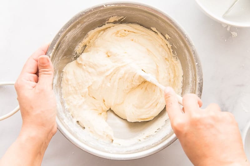 A hand uses a white rubber spatula to finish folding in the flour into batter in a silver bowl.