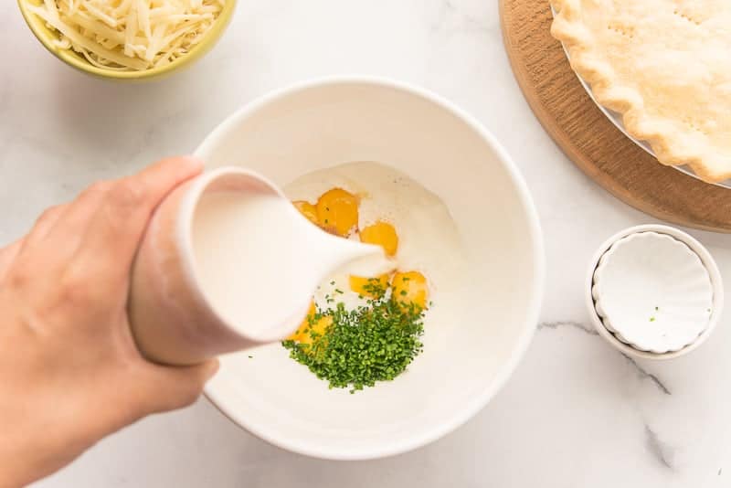 Half-and-half pouring into the white ceramic bowl filled with egg yolks, chives, and spices
