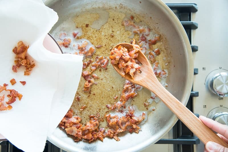 A wooden spoon lifts the cooked bacon pieces from the skillet to a paper towel-lined bowl.