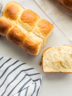 Preview image of Brioche Bread with the heel pulled off to show interior texture of bread. White and blue kitchen towel bottom left. Red and white checkered capped jam jar top left. Corner of another loaf of brioche in top right.