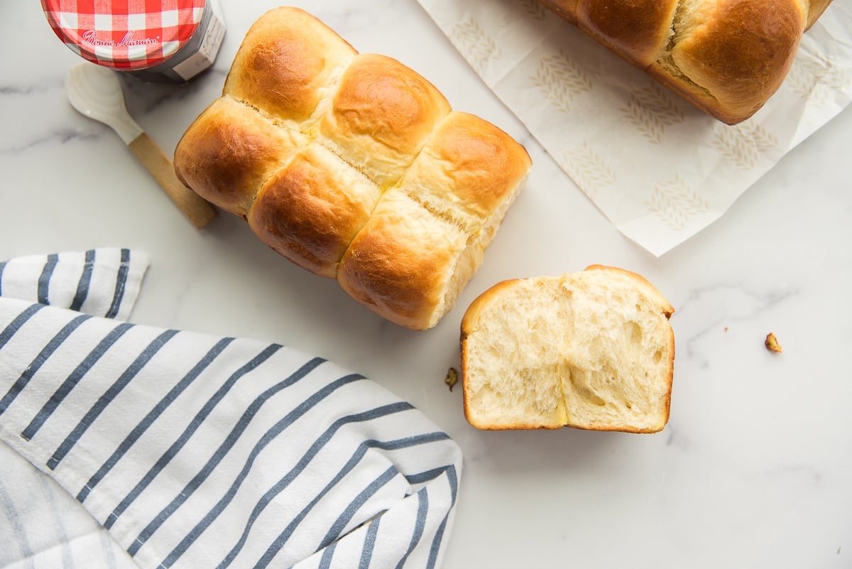 Preview image of Brioche Bread with the heel pulled off to show interior texture of bread. White and blue kitchen towel bottom left. Red and white checkered capped jam jar top left. Corner of another loaf of brioche in top right.