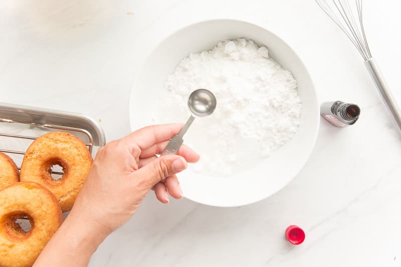 A hand uses a measuring spoon to add extract to a bowl of powdered sugar