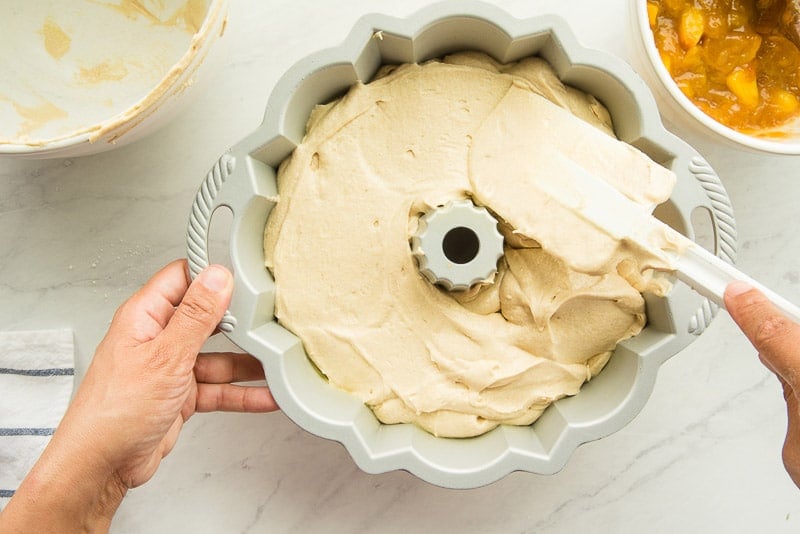 A hand smooths the top of the cake batter in a silver bundt pan using a white rubber spatula.