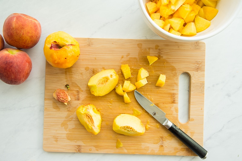 Two peach on a bamboo cutting board. One is peeled and whole, the other is sliced into wedges and diced next to a small paring knife.