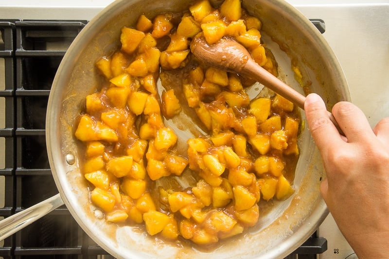 A hand stirs the peach topping in a silver pan with a wooden spoon.