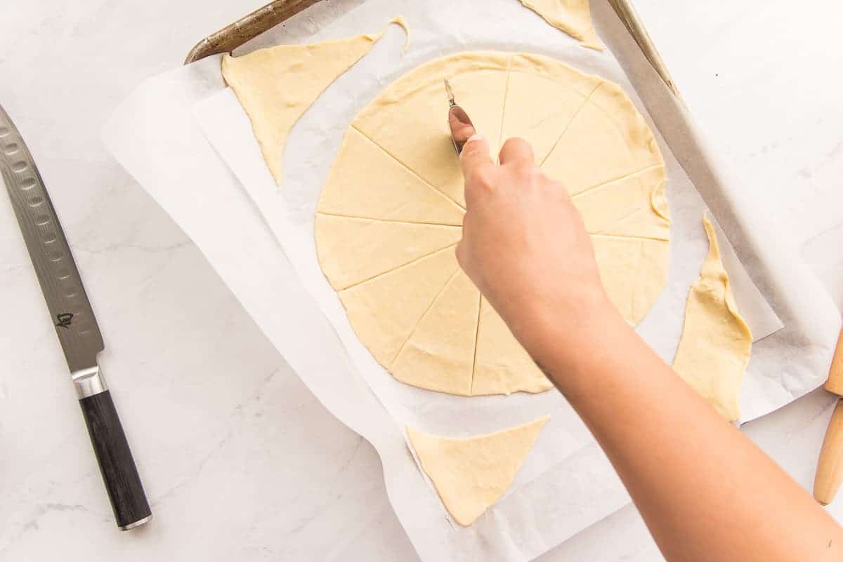 A hand uses a pastry cutter to cut a circle of puff pastry into wedges.