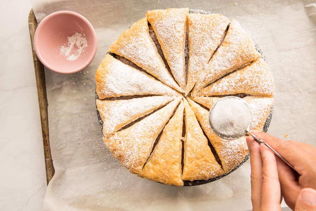 A hand sifts powdered sugar onto the puff pastry topped cheesecake with a tea strainer.